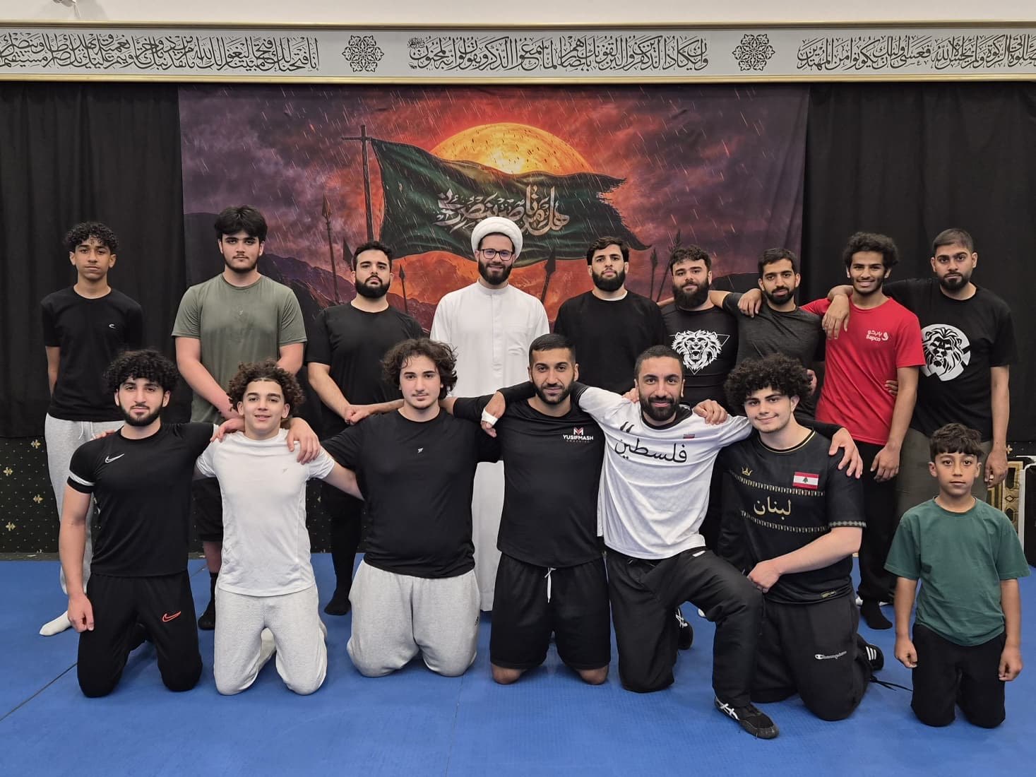 Adults and kids from the club in two rows on the blue mats, arms round each other's shoulders, in front of a painted banner of a green flag against a stormy sky, with a man in a white robe and turban at the centre of the back row.